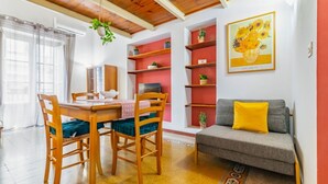 Un comedor con una mesa y sillas de madera, un sofá con un cojín amarillo y un estante rojo con plantas.