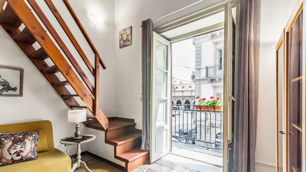 Una escalera conduce a un balcón con vista a la calle, un sofá con un cojín estampado y una mesita con una lámpara.