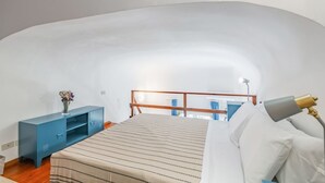 Et soveværelse med skrå væg, seng med hvide sengetøj, en blå kommode og et natbord med lampe.