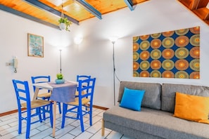 A room with a dining table, blue chairs, a grey sofa with blue and orange cushions, a patterned wall hanging, and a hanging plant.