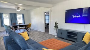 Een moderne woonkamer met een hoekbank, een flatscreen-tv waarop Disney+ te zien is, een eethoek met een tafel en stoelen, en een gang die naar een andere kamer leidt.