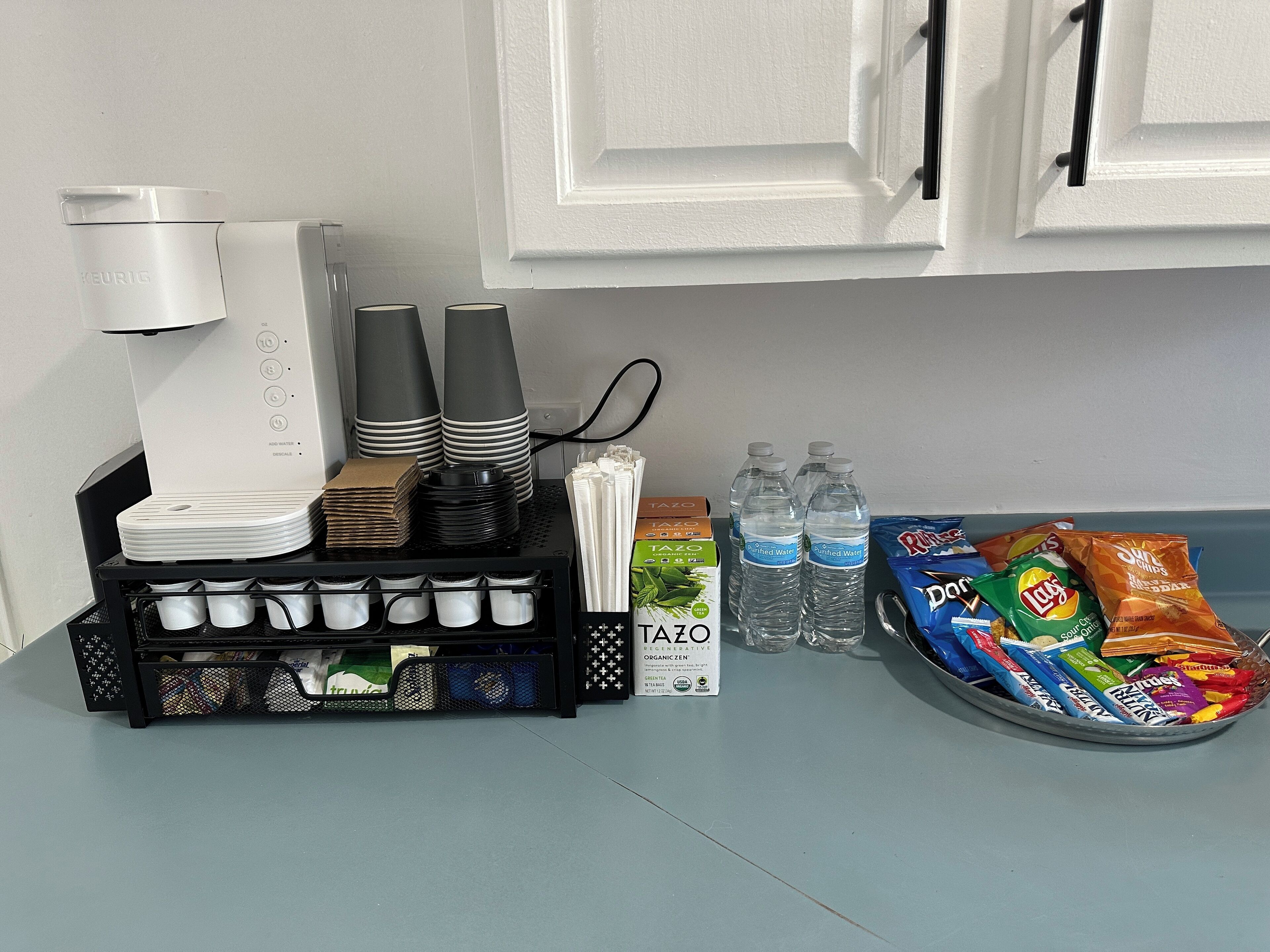 Een keukenhoek met een koffiezetapparaat, gestapelde borden, bekers en een schaal met snacks.