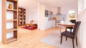 Un salón amplio con una estantería de madera, una mesa de comedor con sillas, una cocina al fondo y un gran ventanal que proporciona luz natural.