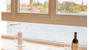 Una habitación con vistas a una iglesia y un río, un radiador, un libro, una vela y una botella de vino en el alféizar de la ventana.