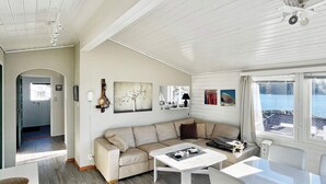 Une pièce moderne et bien éclairée, avec un canapé beige, une table blanche et une vue sur la mer par une grande fenêtre.