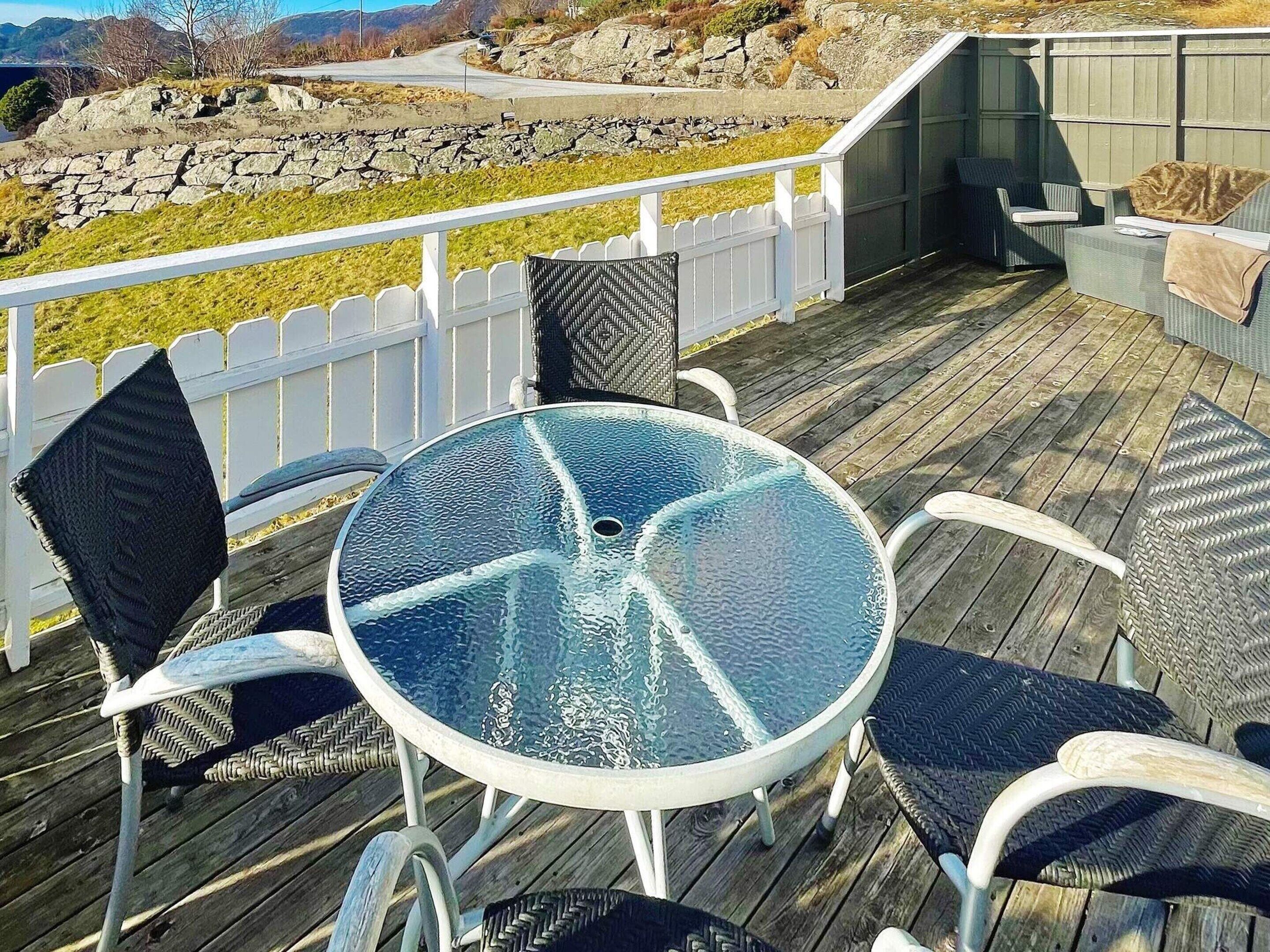 A deck with a round glass table and wicker chairs, overlooking a rocky landscape and a road.