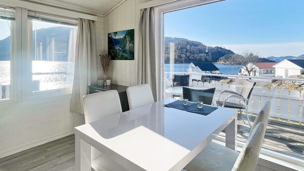 Un espace repas moderne avec une table et des chaises blanches, un tableau au mur et une vue sur un lac et des bâtiments.