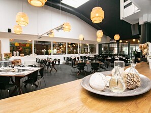 A restaurant interior with multiple tables, hanging pendant lights, and a variety of table settings including plates, glasses, and bottles. - 6 Person Holiday Home on a Holiday Park in Blavand (Blavand)