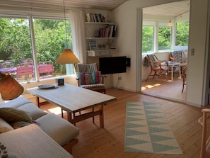 A cozy living room with a sofa, a table, and a TV. There are bookshelves and a window with a view of greenery.