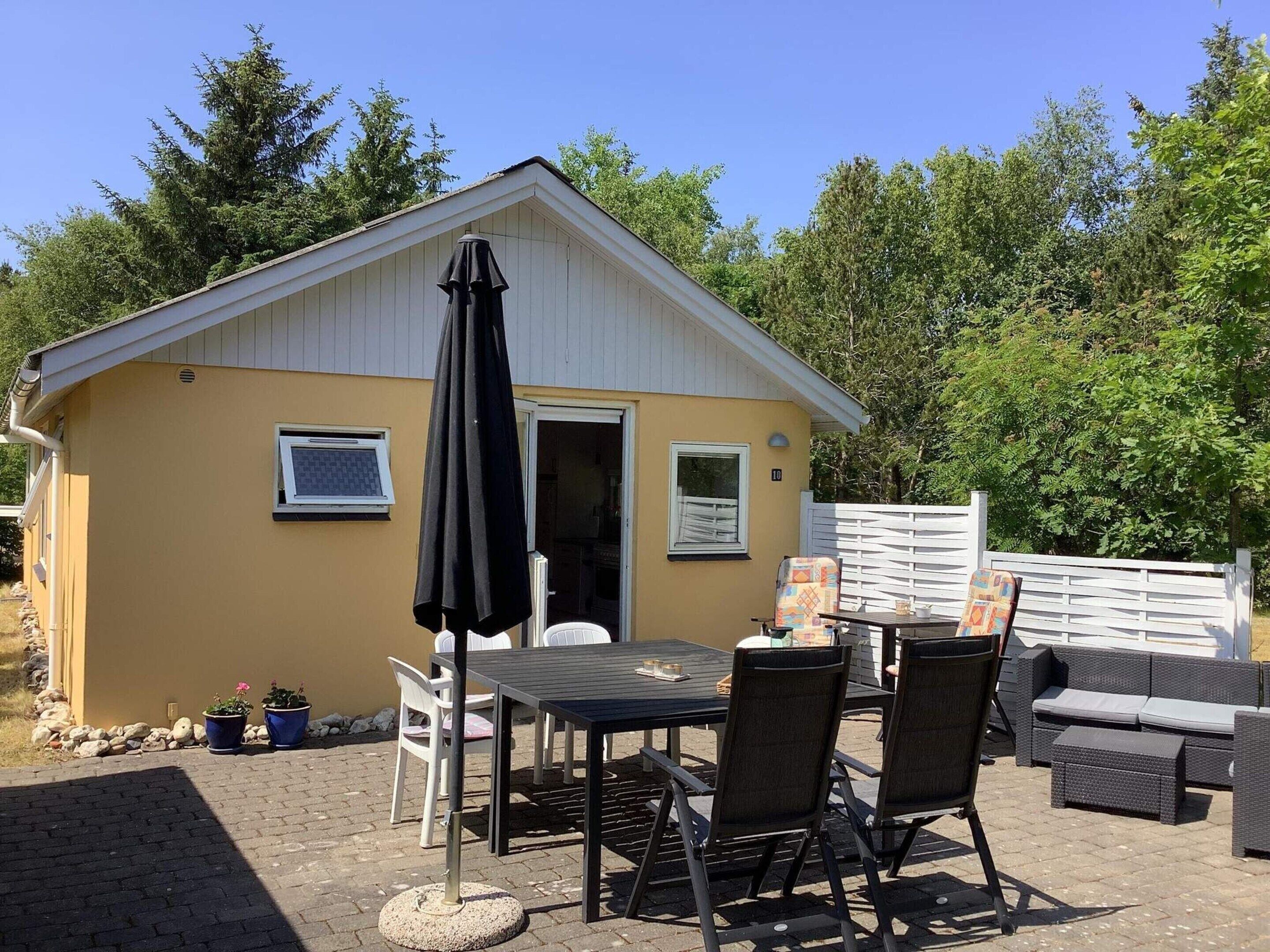 A small, single-story building with a yellow exterior and a white roof. It features a patio with a table and chairs, a black umbrella, and potted plants.