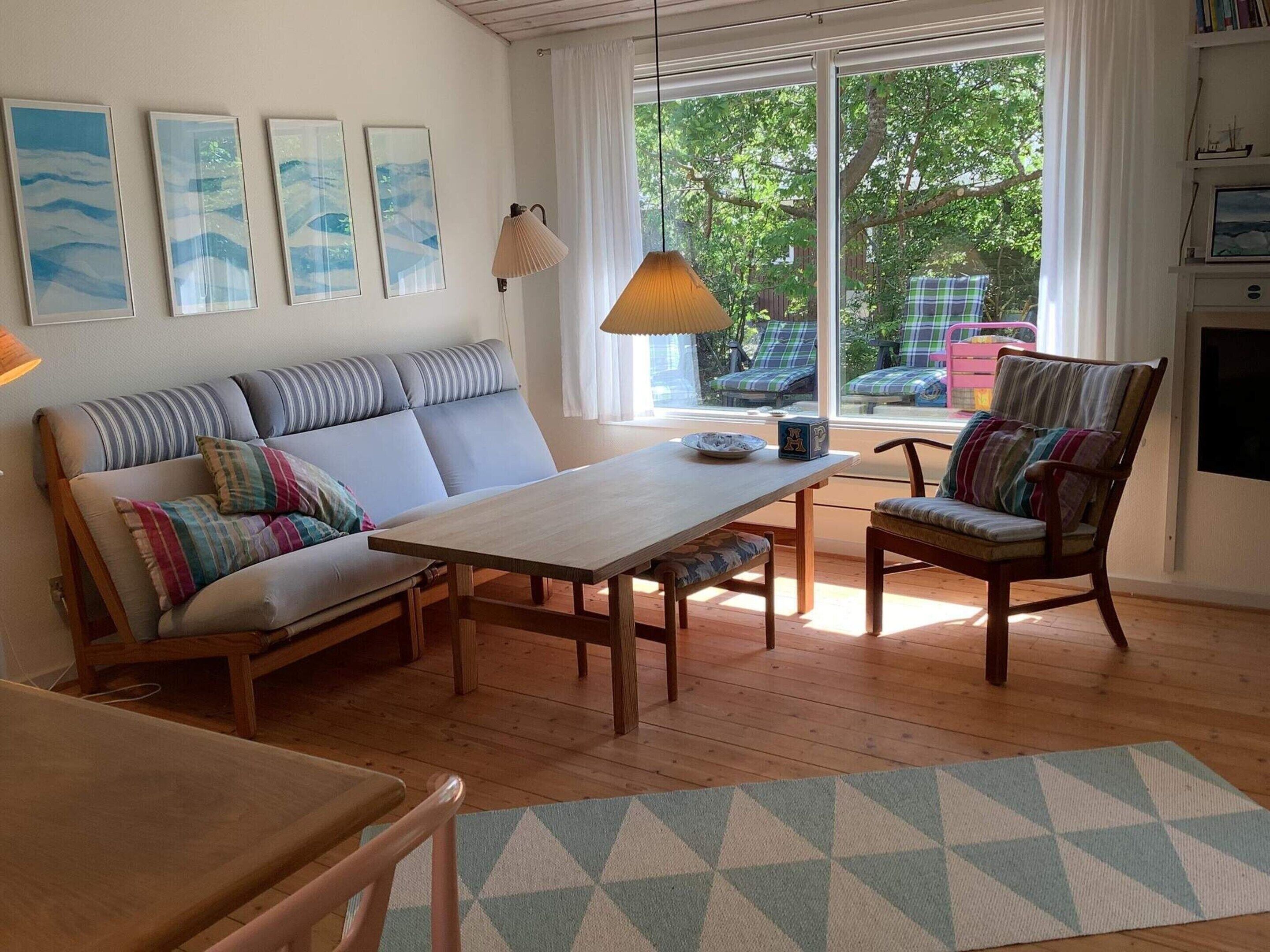 Un salon chaleureux avec un canapé, une table en bois et une chaise. Des tableaux encadrés ornent le mur, ainsi qu’une lampe. De grandes fenêtres offrent une vue sur l’extérieur.