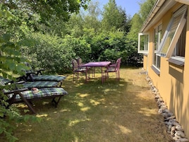 Un patio con mesa y sillas, rodeado por un borde de piedra y abundante vegetación.