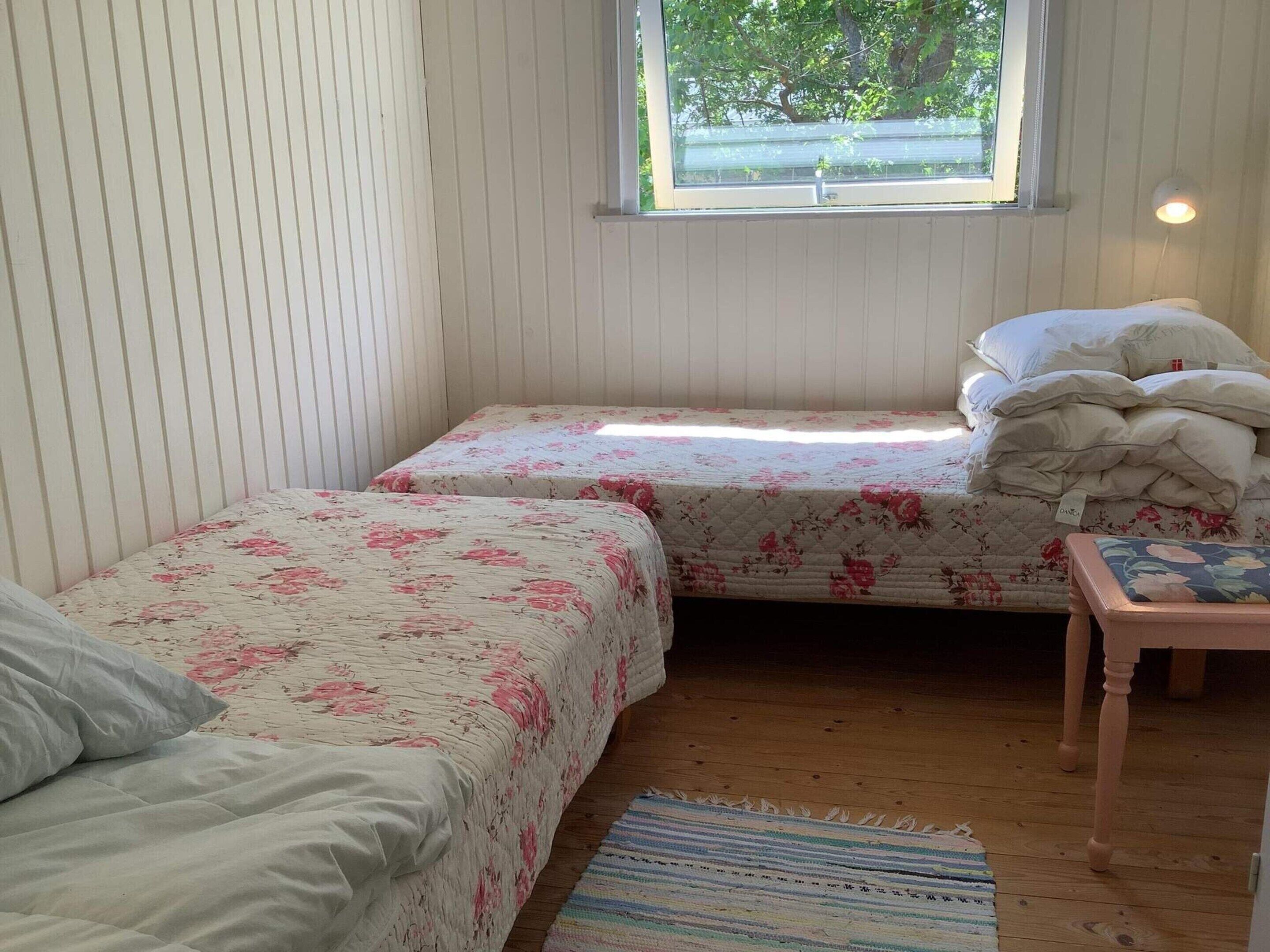 A small, cozy room with two single beds, a wooden floor, a window with a view of trees, and a small table with a cushion.