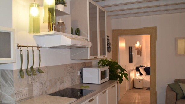Une cuisine moderne avec des meubles blancs, un four à micro-ondes et des ustensiles suspendus. On trouve une plante sur le comptoir et une porte qui donne sur une chambre.