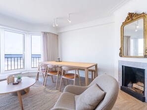 A modern living room with a wooden dining table, comfortable seating, a fireplace, and large windows offering a view of the outdoors. - Bright Apartment With Direct sea View (Middelkerke)