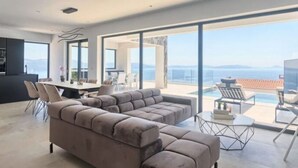 A modern living room with a large sectional sofa, a dining area with a table and chairs, and a balcony with a pool overlooking a scenic view. - Villa Ateneum Comfortable Holiday Residence (Labin)