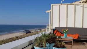 Un balcon agrémenté de plantes en pot, une table ornée d’un vase de fleurs et une vue sur la plage et l’océan.