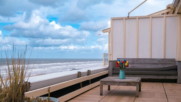Un balcon avec vue sur l’océan, un banc en bois, une petite table et des plantes en pot.