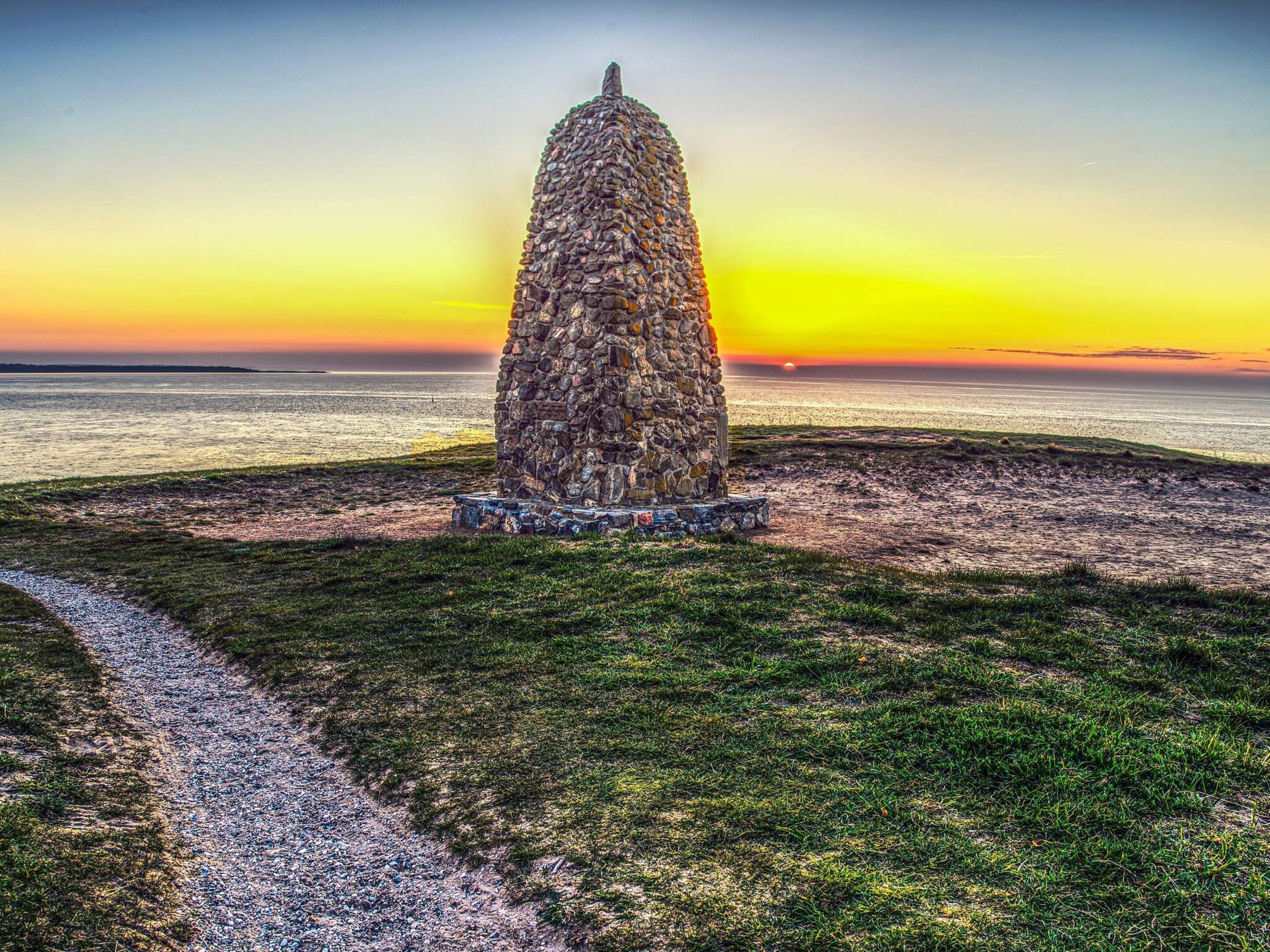 Ett stenmonument med en cirkelformig bas, placerat på en gräsyta med en stig som leder fram till det.