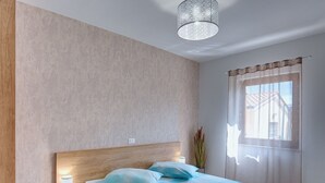A bedroom with a wooden headboard, a bedside table with a lamp, a bed with blue bedding, and a window with sheer curtains. - Apartment in Croatia Near the Coast (Medulin)