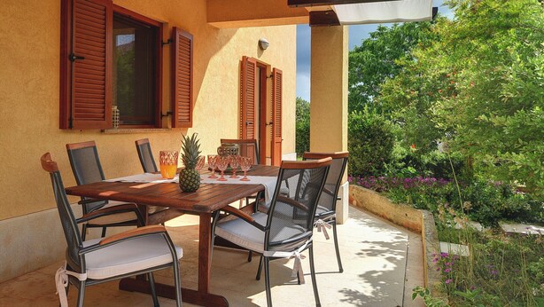 An outdoor patio with a wooden table and chairs, surrounded by a yellow wall and wooden shutters. - Apartment in Croatia Near the Coast (Medulin)