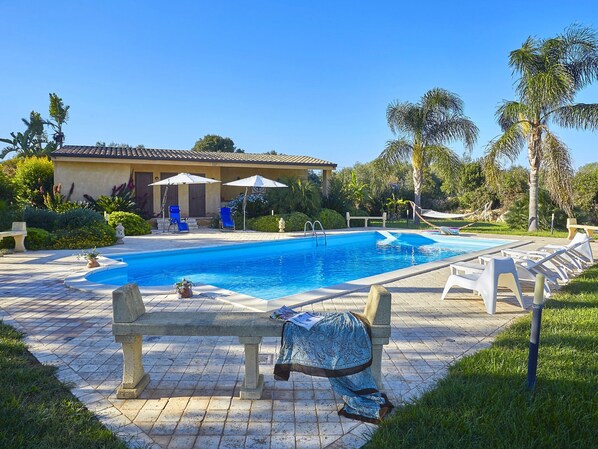 A swimming pool with lounge chairs and umbrellas, surrounded by greenery and a building with a tiled roof.