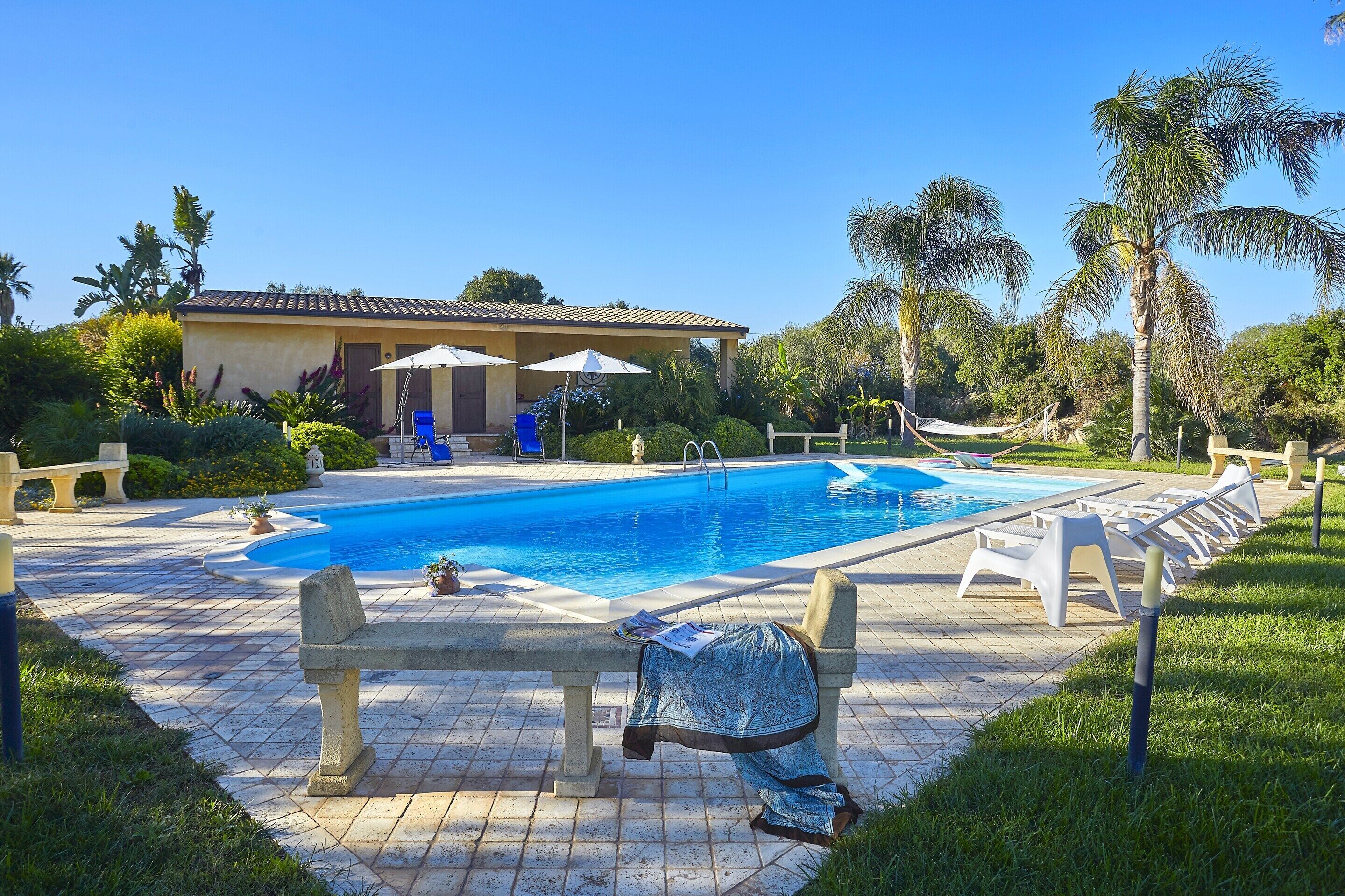 A swimming pool with lounge chairs and umbrellas, surrounded by palm trees and a building with a tiled roof.