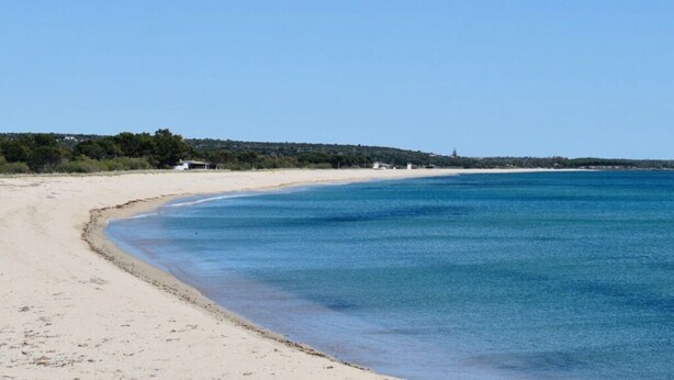 Une plage avec une eau d’un bleu limpide, un rivage sablonneux et quelques bâtiments visibles au loin.