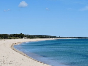 Une plage avec une eau d’un bleu limpide, un rivage sablonneux et quelques bâtiments visibles au loin.