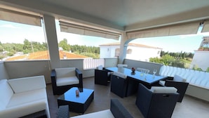 Une terrasse extérieure couverte, avec un canapé blanc, des meubles en rotin noir et une table dressée pour le repas.