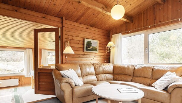 Ein gemütliches Zimmer mit einem ledernen Ecksofa, einem runden Holztisch und großen Fenstern mit Blick ins Freie.