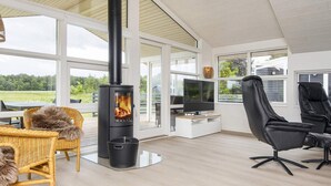 A modern living room with a wood-burning stove, comfortable seating, a flat-screen TV, and large windows offering a view of the outdoors.