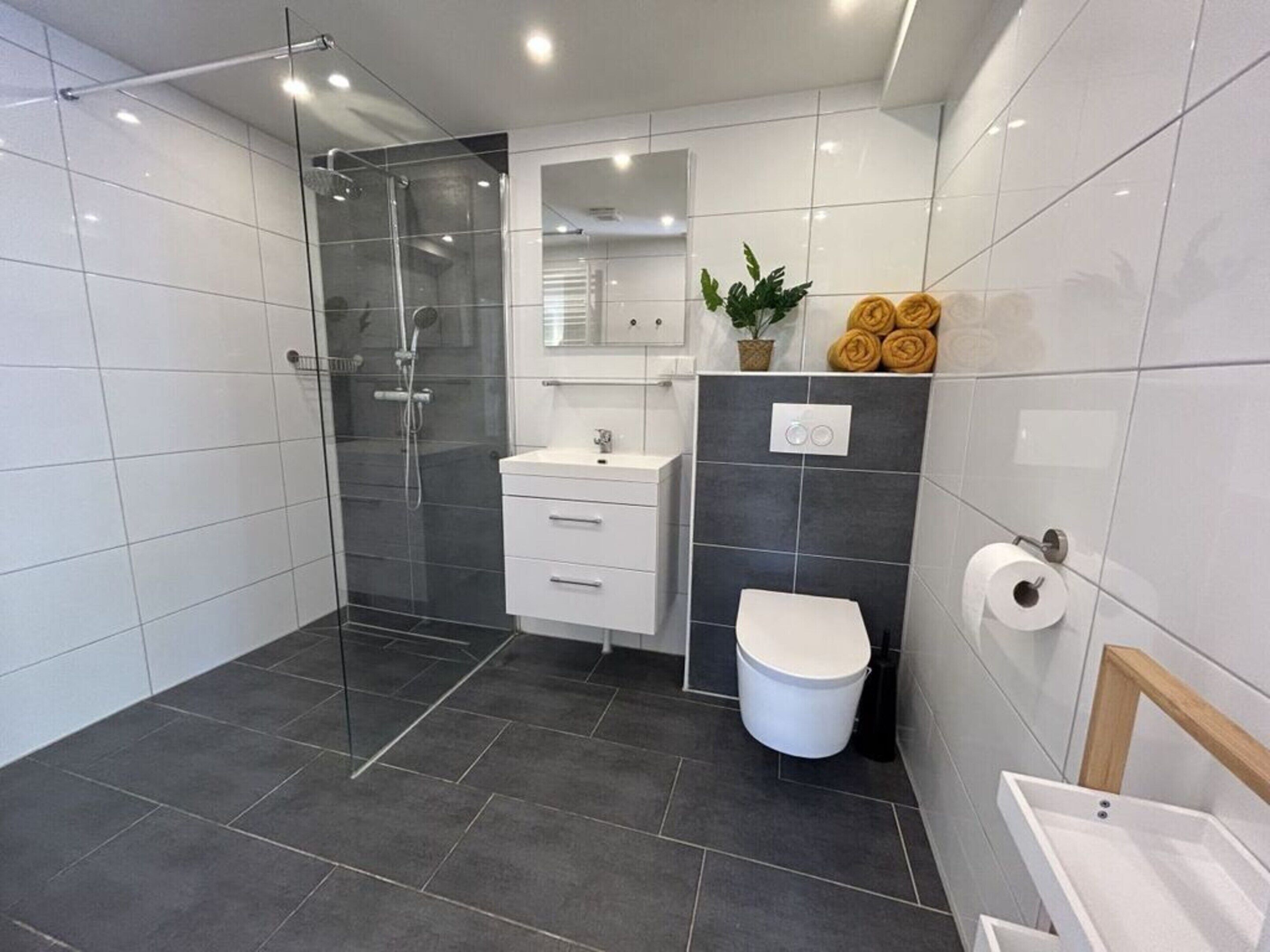 A modern bathroom with a glass-enclosed shower, a white vanity with a mirror, a toilet, and a shelf with towels.