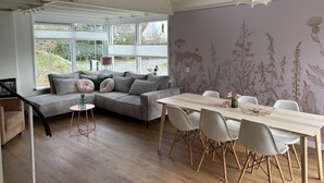Ein modernes Wohnzimmer mit einem grauen Sofa, einem Esstisch mit Stühlen und einem großen Fenster mit Blick auf Grünflächen.