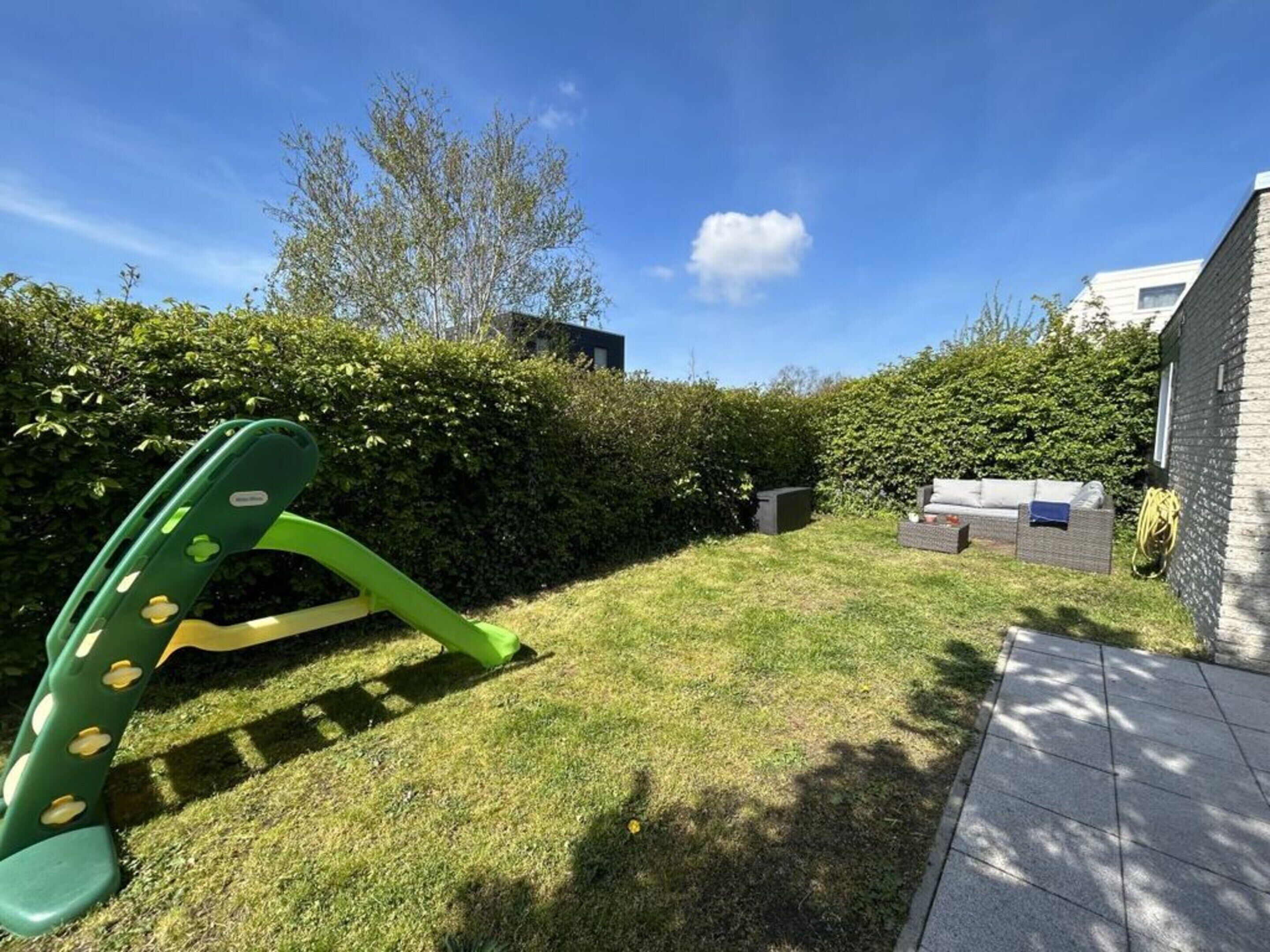 A grassy backyard with a green play structure, a hedge, and a brick wall.