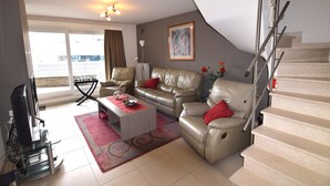 A living room with a sofa, coffee table, and a TV. There is a staircase and a window with a view. - Apartment in a Central Location (Blankenberge)