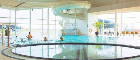 Une piscine intérieure avec un toboggan, de grandes fenêtres et un espace détente avec des fauteuils.