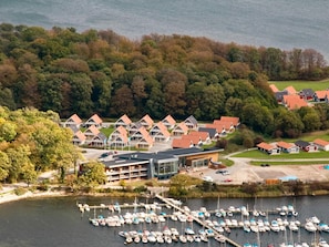 Ein Seehotel mit mehreren Gebäuden, einer Marina mit zahlreichen Booten und einer bewaldeten Hügelkuppe im Hintergrund.