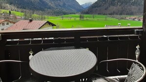 A balcony with a table and chairs overlooking a valley with green fields and mountains.