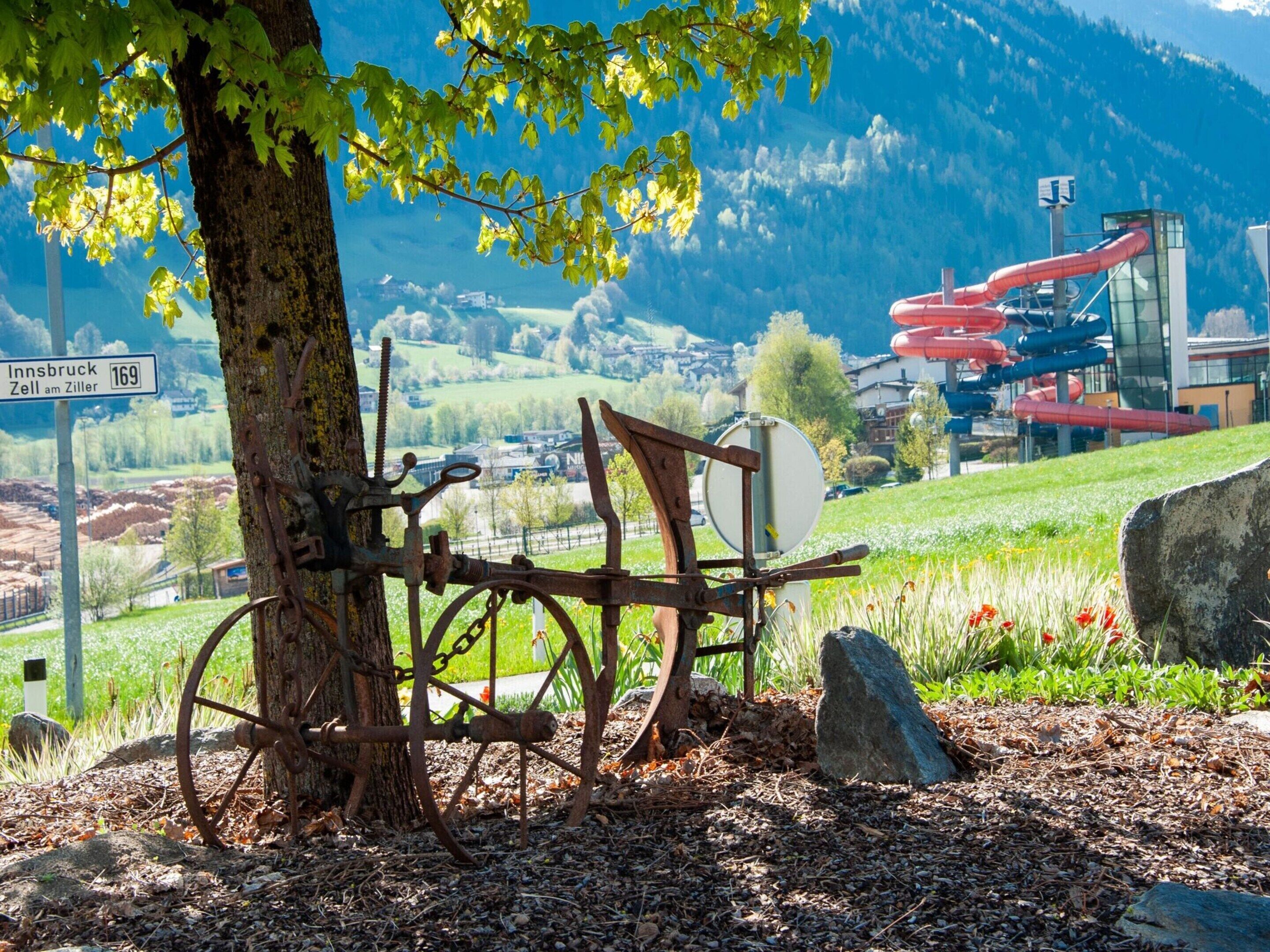 A rustic metal structure with a wheel and chains, set against a backdrop of a mountain landscape and a modern building with red pipes.