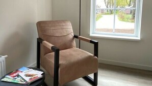 Une pièce comprenant un seul fauteuil marron, une petite table ronde avec des magazines, un lampadaire et une fenêtre donnant sur des arbres et des maisons.