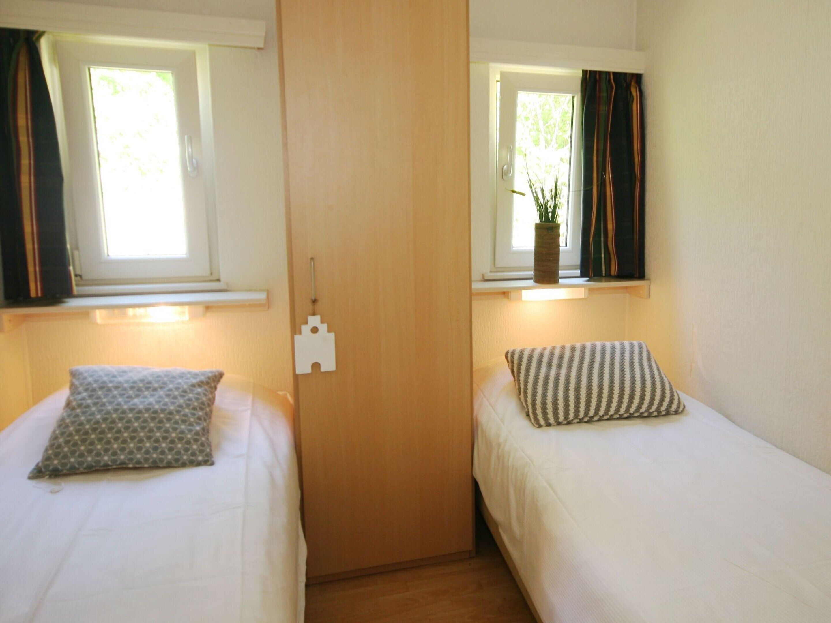 A room with two single beds, each with a pillow and a window. There is a wooden door in the center, and a small plant on the windowsill of the right bed.