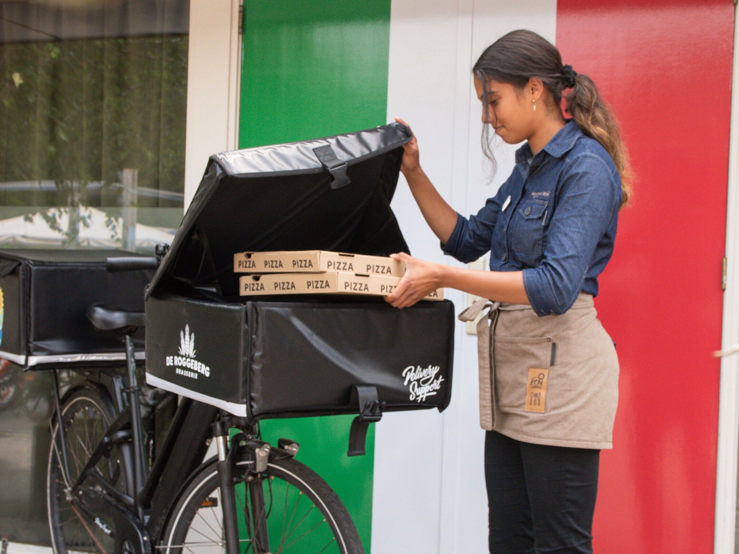 A delivery person loading pizza boxes into a black delivery bike with a De Roster logo.