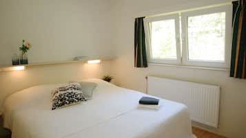 A neatly made bed with a white comforter and a patterned pillow, a bedside table with a book and a vase with flowers, a window with curtains, and a radiator.