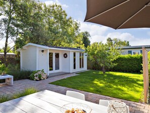 A white wooden shed with a door and windows, a brick patio, and a lawn. - Lovely, Fully Furnished Chalet for 4 People (Goedereede)