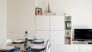 Un comedor con mesa de madera dispuesta para cuatro personas, sillas blancas y un armario blanco con televisor y estanterías.