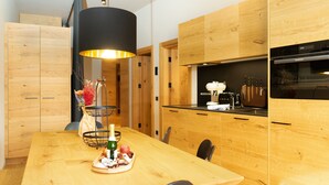 A modern kitchen with wooden cabinetry, a black pendant light, and a dining table set with a bottle, apple, and decorative items. - Apartment in Schrocken Near Ski Arlberg (Schroecken)