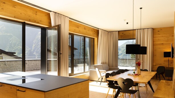 A modern kitchen with a large island, a dining area with a wooden table and chairs, and a large window offering a view of the mountains. - Apartment in Schrocken Near Ski Arlberg (Schroecken)
