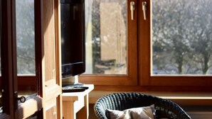 A room with a television on a wooden stand, a wicker chair, and a view of trees through a large window. - Deimann Modern Retreat (Schmallenberg)