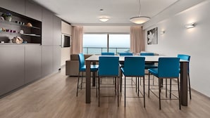 Un espace repas moderne avec une grande table et des chaises bleues, une cuisine aux meubles gris et une vue sur la mer grâce à de grandes fenêtres.